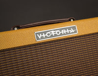A close up of the black and silver Victoria logo plate on the front of a tweed Vicky Verb model amp. 